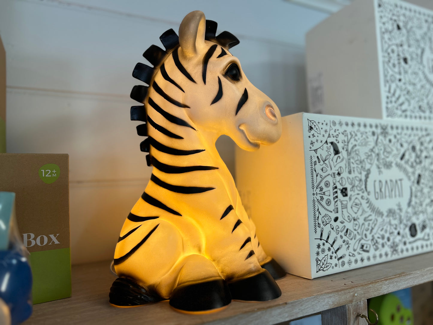 Zebra shaped nightlight facing to the right.. The light is on and shows a warm yellow glow. The Zebra is in a sitting position.