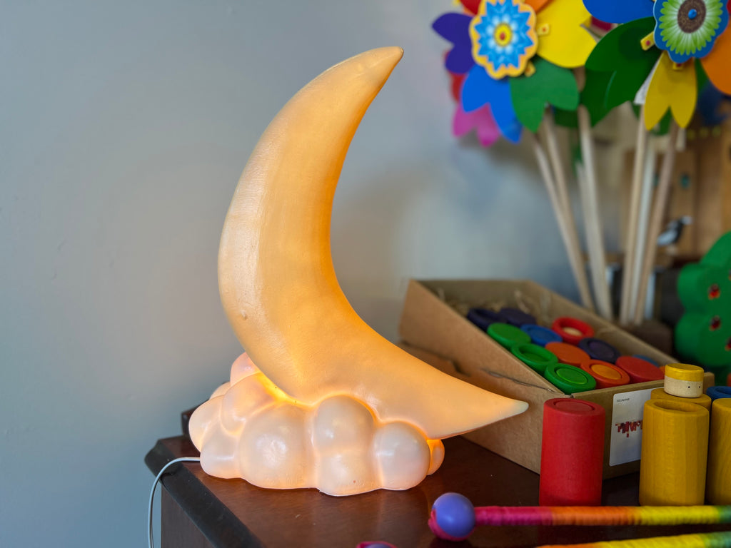 A night light in the shape of a crescent moon sits on a small cloud shape.  The lightis on and gives off a warm yellow glow.  There are colourful children's toys in the background.